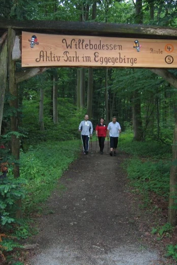 Drei Personen spazieren einen Waldweg entlang zum Aktiv-Park im Eggegebirge, umgeben von Bäumen.