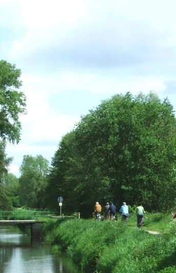 Gruppe von Radfahrern auf einem naturnahen Weg entlang eines Kanals unter blauem Himmel.