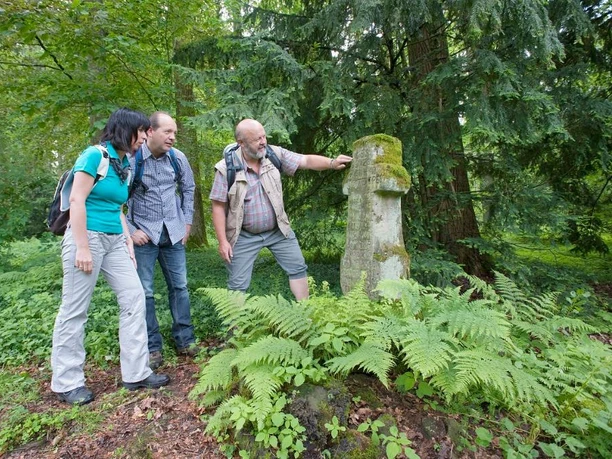 Pestfriedhof Leiberg Drei Menschen stehen im Wald neben einer alten Grabstele, umgeben von Farnen und Bäumen.