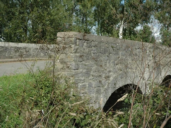 Brücke Steinbrücke mit zwei Bögen, umgeben von grünem Wiesenrand und hohen Bäumen im Hintergrund.