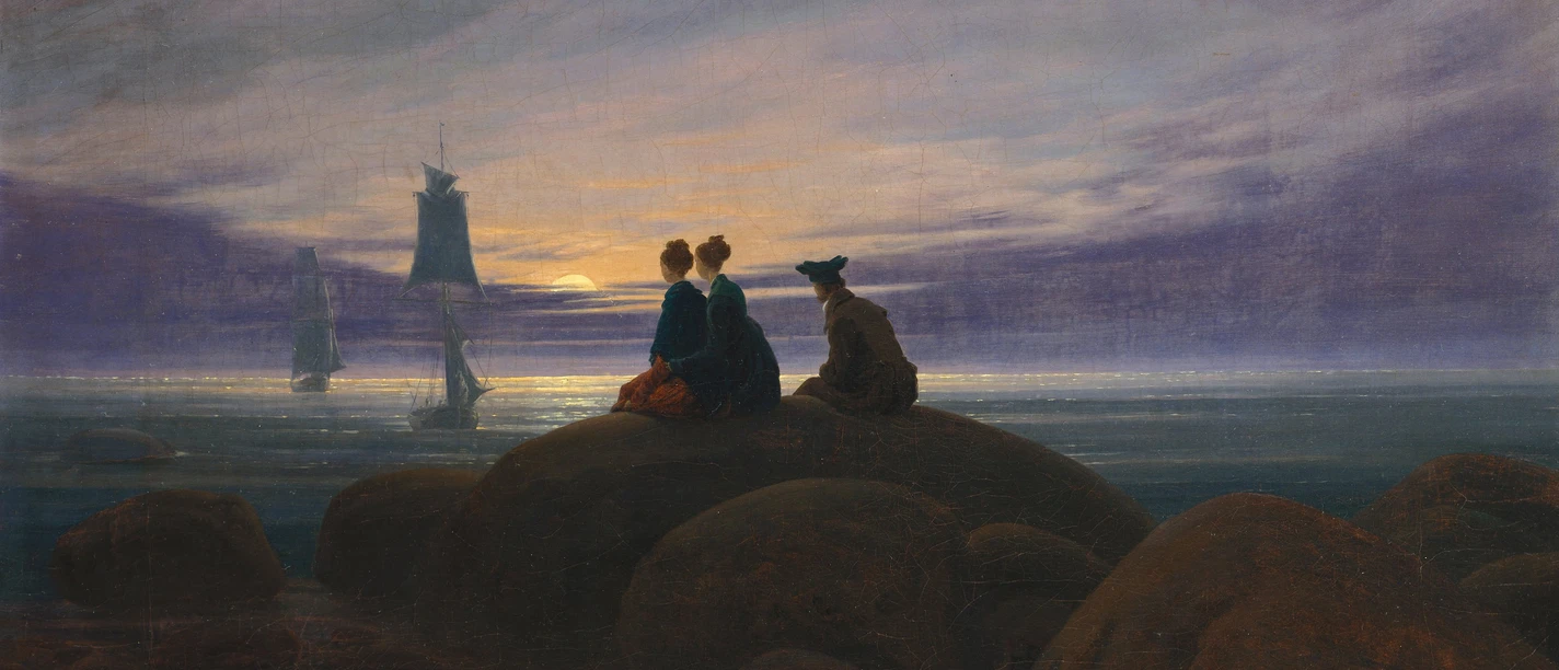 Auf dem Bild sind große Felsen am Meer zu sehen, auf denen drei Personen von hinten erkennbar sind. Im Hintergrud sieht man Segelboote.