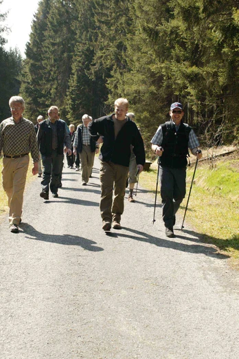 Eine Gruppe älterer Männer wandert fröhlich auf einem Waldweg, umgeben von hohen Bäumen.