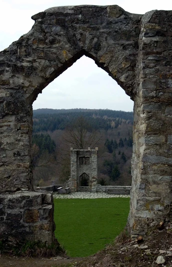 Steinerne Ruinen mit einem Torbogen, durch den ein weiteres Burggemäuer und Wald zu sehen sind.