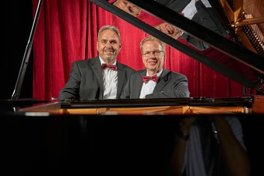 Duo_Kurzweil_230707_0632 (1) Zwei Männer in Anzügen mit Fliegen sitzen lächelnd vor einem Klavier. Im Hintergrund sind rote Vorhänge sichtbar.