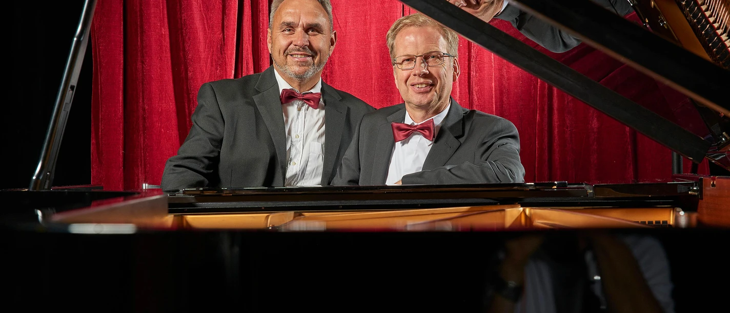 Duo_Kurzweil_230707_0632 (1) Zwei Männer in Anzügen mit Fliegen sitzen lächelnd vor einem Klavier. Im Hintergrund sind rote Vorhänge sichtbar.