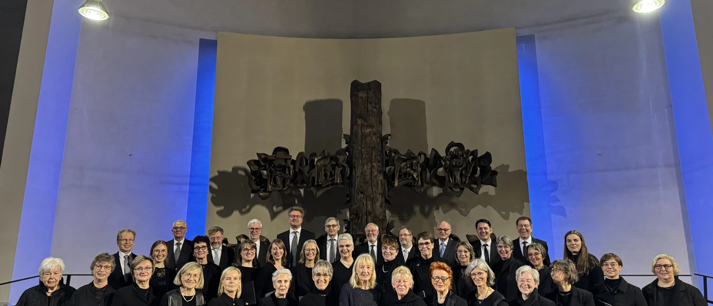 Chorfoto musica vocalis November 2025 Eine große Gruppe von Menschen steht auf einer Bühne in einem Kirchenraum. Sie tragen schwarze Kleidung und halten Ordner in den Händen, während ein kunstvolles Kreuz im Hintergrund beleuchtet ist.