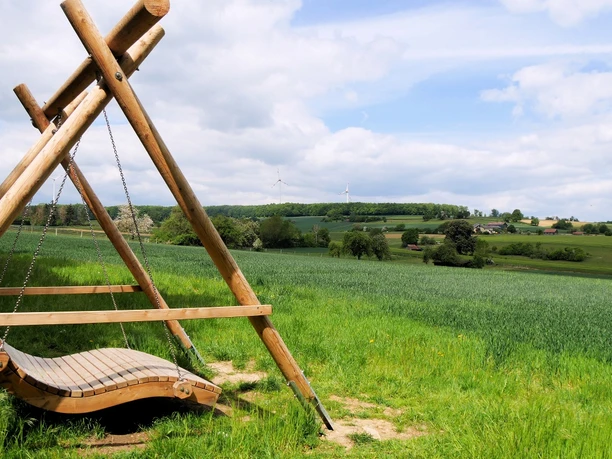 Landschaftsschaukel bei Bellersen Eine Holzschaukel bietet Ausblick auf grüne Felder, sanfte Hügel und Windräder unter blauem Himmel.