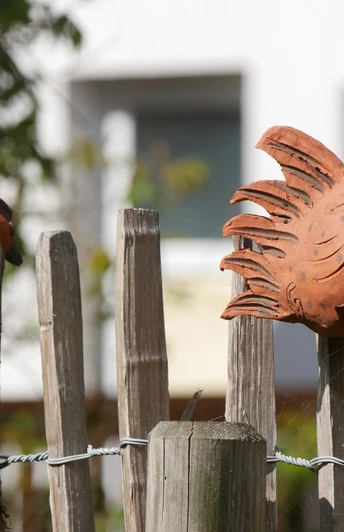 Zaundeko in Bellersen Terrakotta-Hühner auf Holzzaun in Bellersen, umgeben von grünen Pflanzen und unscharfem Hintergrund.