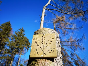 Ein hölzernes Wegezeichen mit der Aufschrift "AW" ist an einem Baum vor klarem blauen Himmel angebracht.