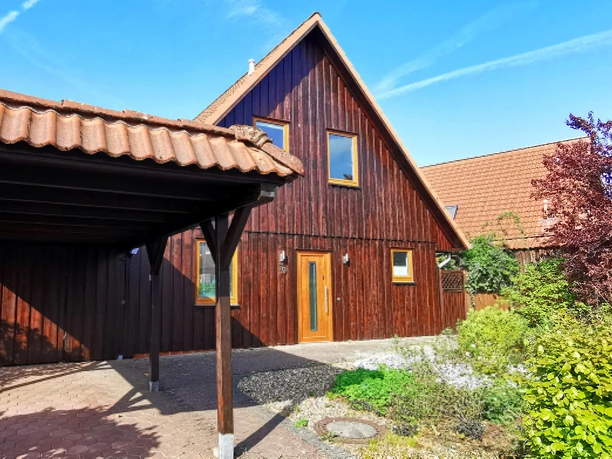 Aussenansicht mit Eingagstür Ein charmantes Holzhaus mit einem steilen, gedeckten Dach. Der Garten ist grün und einladend, und der Himmel ist klar und blau.