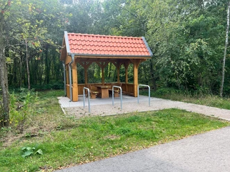 Holz-Schutzhütte mit rotem Ziegeldach, Sitzgruppe und Fahrradbügeln an der Fahrradstraße.