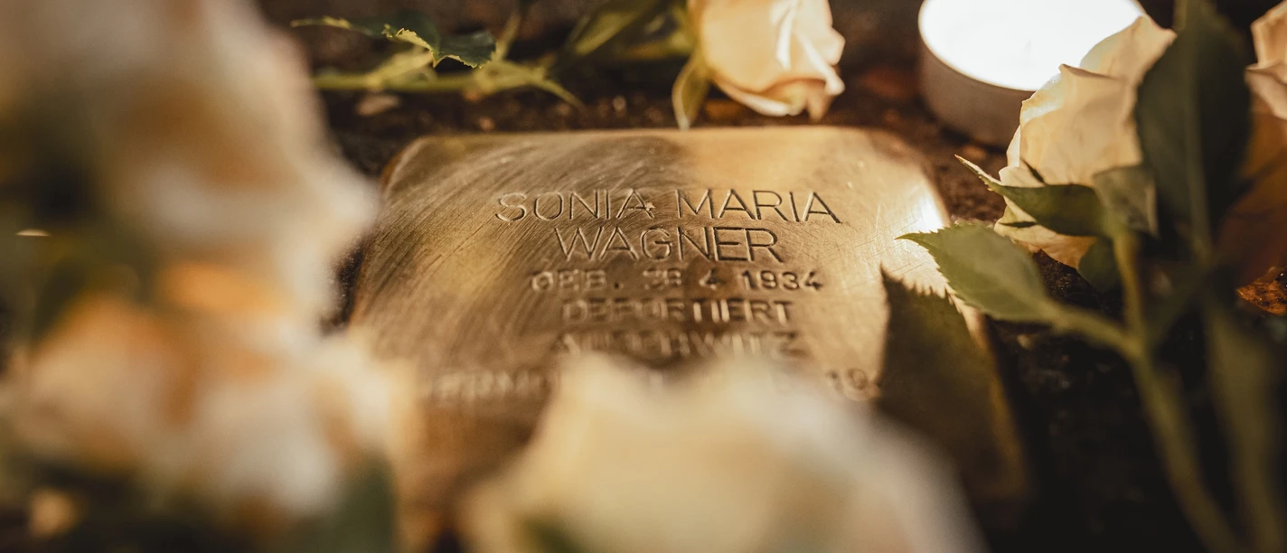 Nahaufnahme von einem Stolperstein mit weißen Rosen und einer Kerze