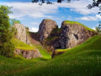 Grüner Hügel mit steinernen Erhebungen und Wiese unter blauem Himmel mit Wolken, umgeben von Bäumen.