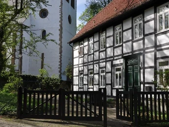 Historisches Fachwerk-Pfarrhaus neben einer Kirche, umgeben von Bäumen und mit Holzzaun im Vordergrund.