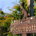 Schild Wanderweg "Hasenpatt" Wegweiser "Hasenpatt" aus Holz mit geschnitztem Hasenmotiv unter umgebenden grünen Büschen.