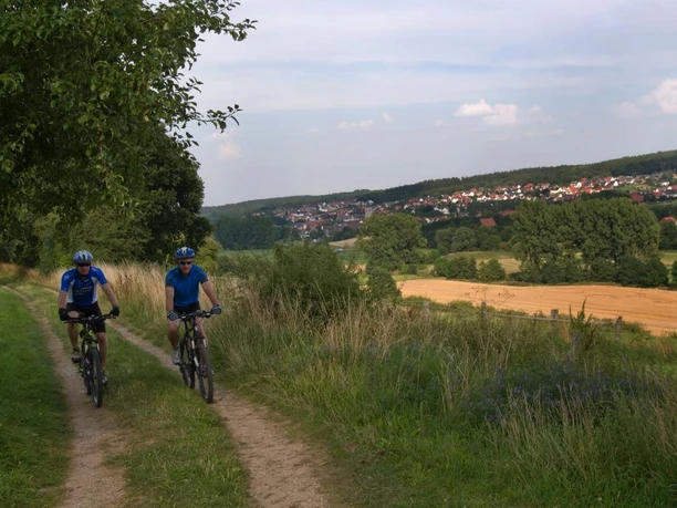 Naturerlebnis im Almetal Zwei Radfahrer auf einem Feldweg durch eine weite, grüne Landschaft mit Blick auf ein Dorf.