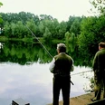 Zwei Männer stehen am Ufer eines üppig bewachsenen Sees, vertieft ins Fischen bei ruhigem Wetter.