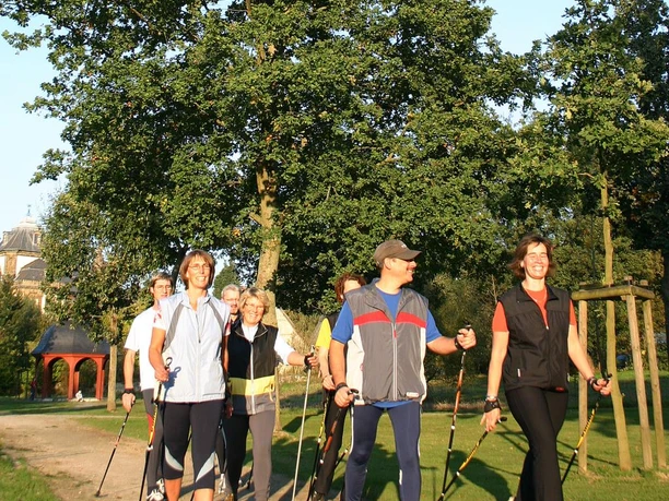Sechs Menschen beim Nordic Walking auf einem Parkweg, umgeben von grüner Natur und Bäumen.