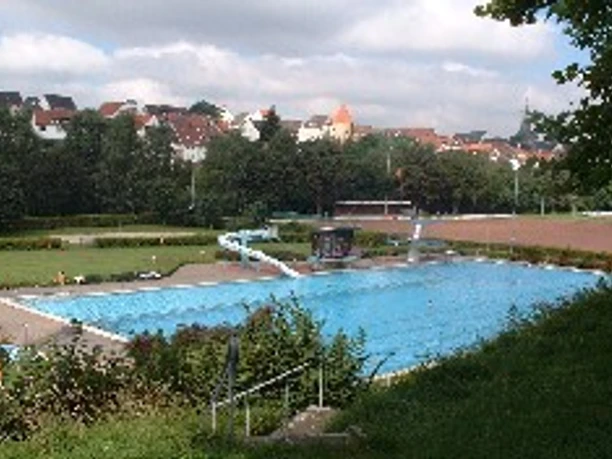 Freibad mit weitläufigem Becken, umgeben von Bäumen und angrenzendem Sportplatz, Siedlung im Hintergrund.