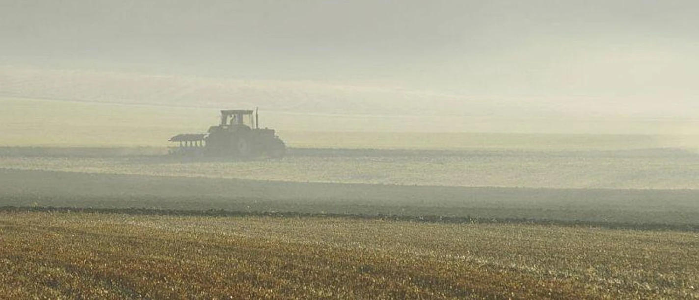 Ein Traktor pflügt ein weites Feld unter bewölktem Himmel, in einer ländlichen und ruhigen Umgebung.