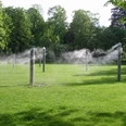 Nebelwiese Bad Lippspringe Sechs hohe Stelen sprühen feinen Wassernebel auf einer grünen Wiese, umgeben von Bäumen.
