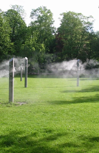 Nebelwiese Bad Lippspringe Sechs hohe Stelen sprühen feinen Wassernebel auf einer grünen Wiese, umgeben von Bäumen.