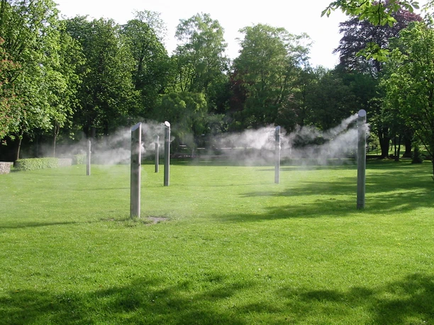 Nebelwiese Bad Lippspringe Sechs hohe Stelen sprühen feinen Wassernebel auf einer grünen Wiese, umgeben von Bäumen.