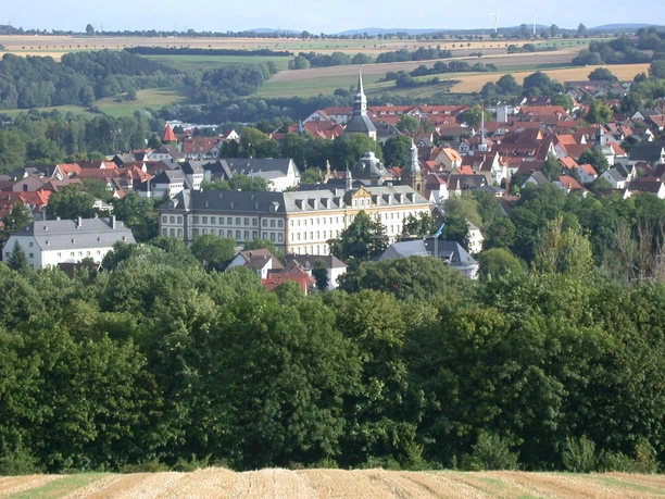Barockes Schloss mit Park in grüner Landschaft, umgeben von einem malerischen Dorf mit Kirchturm.