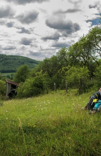 Ein Wanderpaar sitzt auf einer Wiese mit Rucksäcken, umgeben von hügeliger Landschaft und Wolkenhimmel.