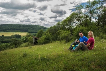 Wanderpaar bei der Rast Ein Wanderpaar sitzt auf einer Wiese mit Rucksäcken, umgeben von hügeliger Landschaft und Wolkenhimmel.