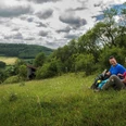 Wanderpaar bei der Rast Ein Wanderpaar sitzt auf einer Wiese mit Rucksäcken, umgeben von hügeliger Landschaft und Wolkenhimmel.