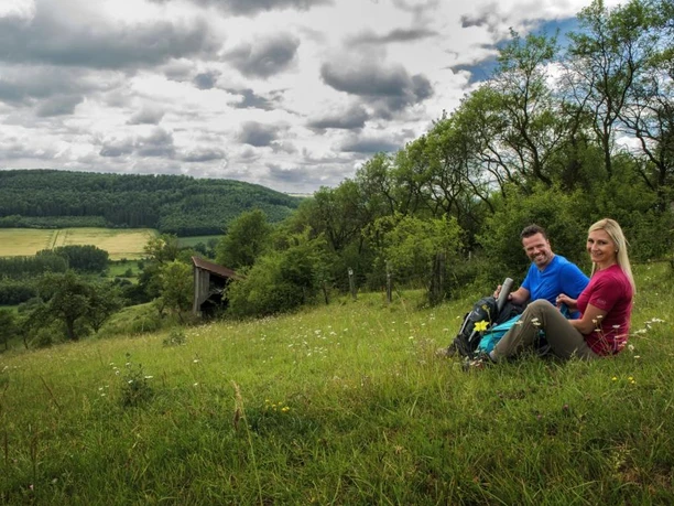 Wanderpaar bei der Rast Ein Wanderpaar sitzt auf einer Wiese mit Rucksäcken, umgeben von hügeliger Landschaft und Wolkenhimmel.