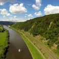 Weserhoehen_Weserskywalk Luftaufnahme eines Flusses, umrahmt von grünen Hügeln und Bäumen, mit einem Boot und blauen Himmel.