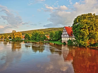 Fachwerkhaus am Flussufer der Weser, umgeben von üppigem Grün unter einem blauen Himmel.