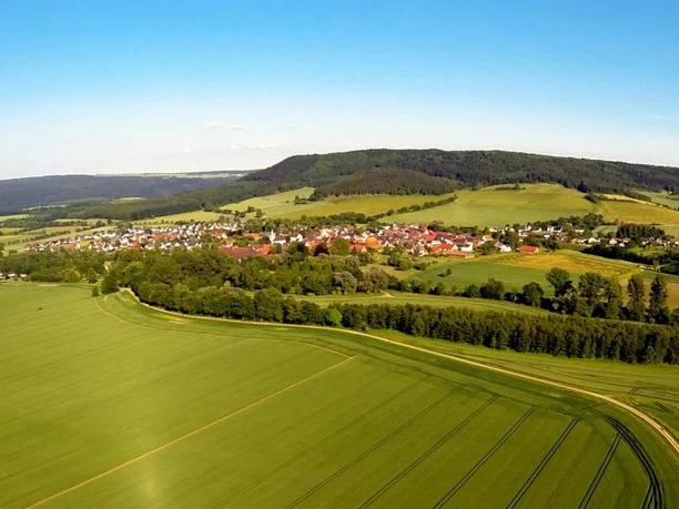 Blick auf Amelunxen im Nethetal Luftaufnahme von Amelunxen, einem Dorf umgeben von grĂĽnen Feldern und sanften HĂĽgeln im Nethetal.
