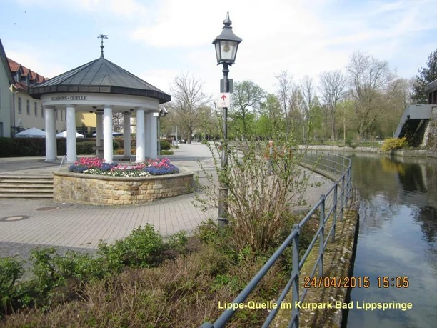 Bad Lippspringe - Lippe-Quelle Pavillon bei der Lippe-Quelle, umgeben von blühenden Blumen und nahe einem Wasserlauf im Kurpark.