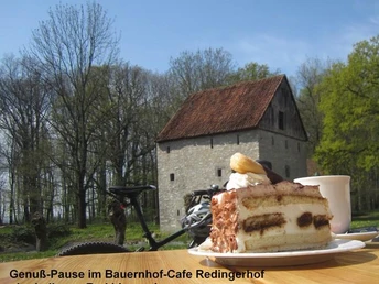 Zwei Stück Kuchen und eine Tasse Kaffee auf einem Tisch, mit einem Fahrrad im Hintergrund, umgeben von einem historischen Gebäude inmitten einer Waldlandschaft.