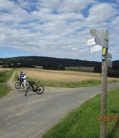 Blick auf den Steinweg von Süd-Westen Ein Radweg in ländlicher Landschaft; Wegweiser; zwei Fahrräder und Personen auf einem asphaltierten Weg.