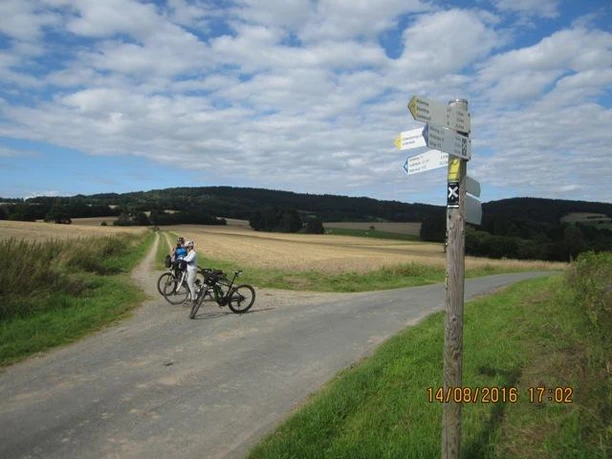 Blick auf den Steinweg von Süd-Westen Ein Radweg in ländlicher Landschaft; Wegweiser; zwei Fahrräder und Personen auf einem asphaltierten Weg.