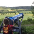 Ein Kind betrachtet die weite Landschaft durch ein großes, blaues Sphaeriskop in ländlicher Umgebung.