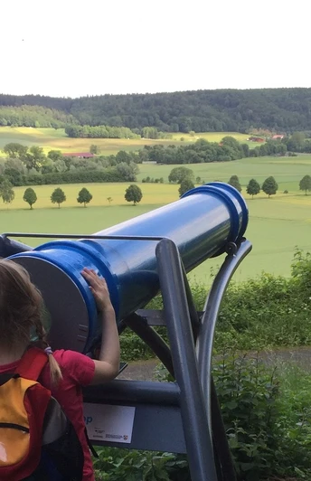 Ein Kind betrachtet die weite Landschaft durch ein großes, blaues Sphaeriskop in ländlicher Umgebung.