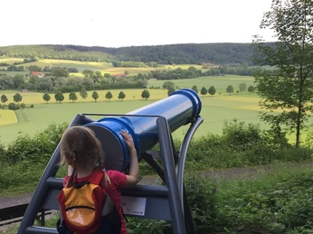 Ein Kind betrachtet die weite Landschaft durch ein großes, blaues Sphaeriskop in ländlicher Umgebung.