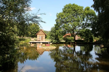 Backhaus am Dorfteich Backhaus an einem ruhigen Dorfteich, umgeben von Bäumen und Wiesen, spiegelt im klaren Wasser.