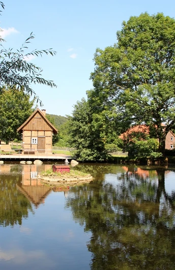 Backhaus am Dorfteich Backhaus an einem ruhigen Dorfteich, umgeben von Bäumen und Wiesen, spiegelt im klaren Wasser.