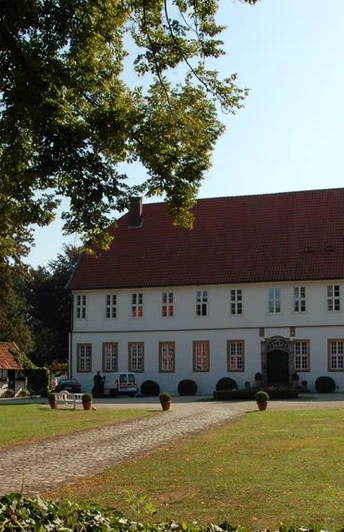 Historischer Gutshof mit rotem Dach, flankiert von Fachwerkgebäuden, umgeben von grüner Natur.