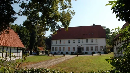 Gut Hiddenhausen Historischer Gutshof mit rotem Dach, flankiert von Fachwerkgebäuden, umgeben von grüner Natur.