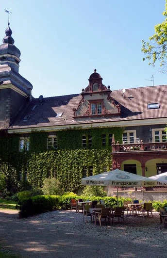 Schloss Mühlenburg mit Efeu bewachsen, Turm mit Uhr, Terrasse mit Tischen und Sonnenschirmen.