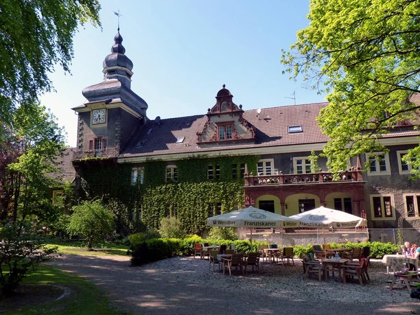 Schloss Mühlenburg Schloss Mühlenburg mit Efeu bewachsen, Turm mit Uhr, Terrasse mit Tischen und Sonnenschirmen.