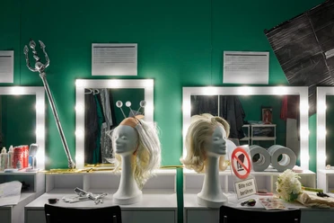Lighted vanity tables with wig heads and props in front of a green wall