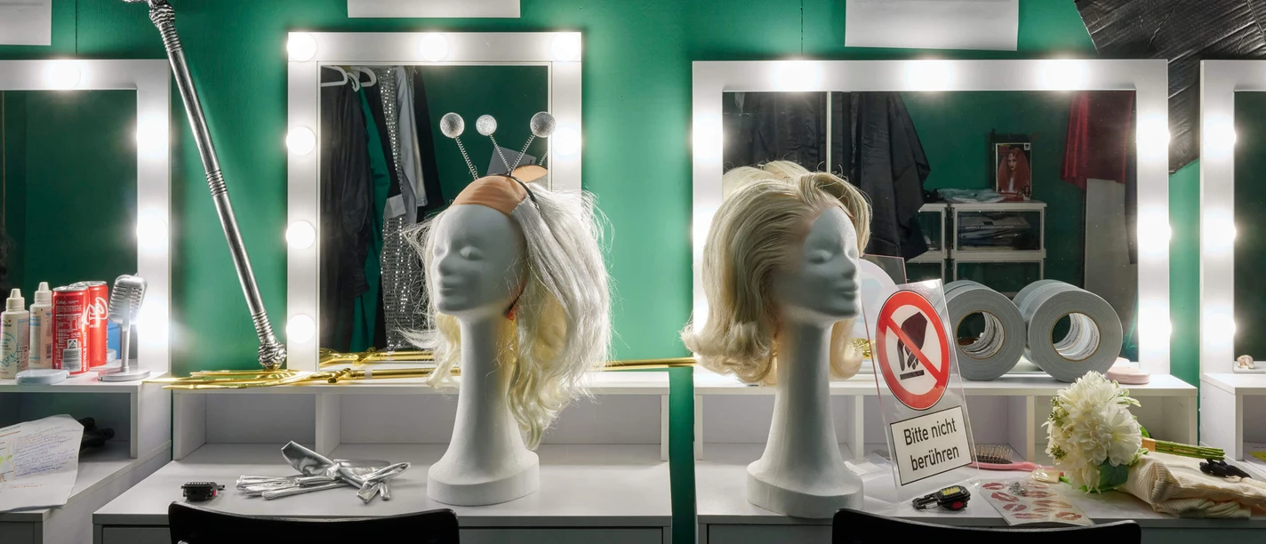Lighted vanity tables with wig heads and props in front of a green wall